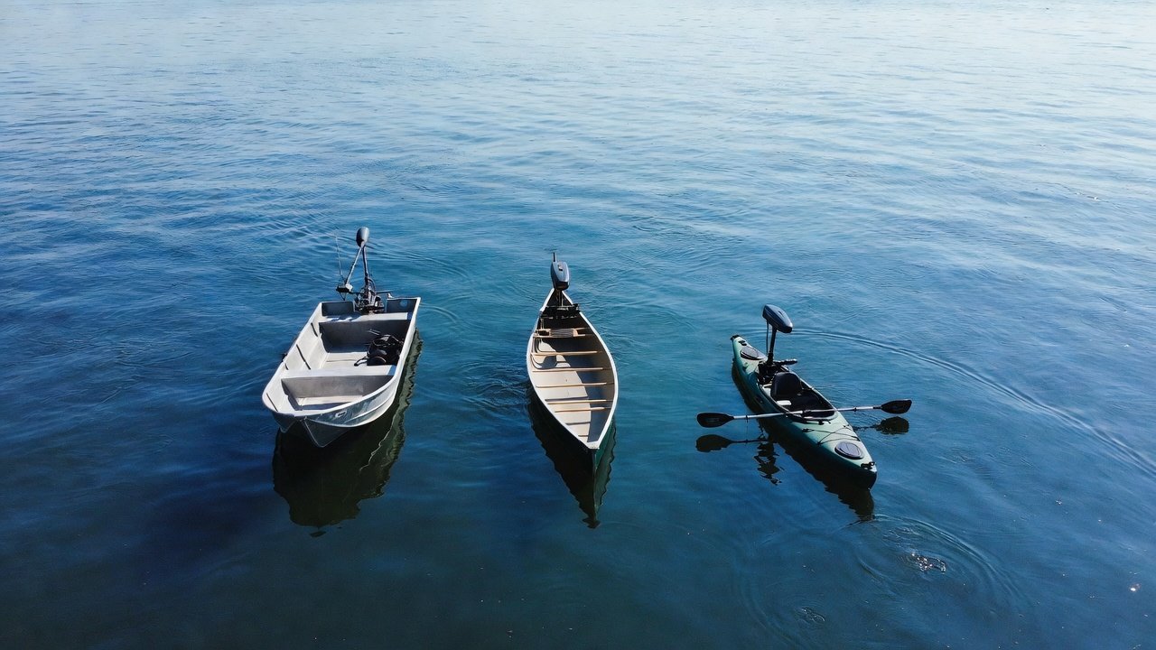 Overhead view of a jon boat, canoe, and fishing kayak on calm blue freshwater water, each fitted with a small electric trolling motor
