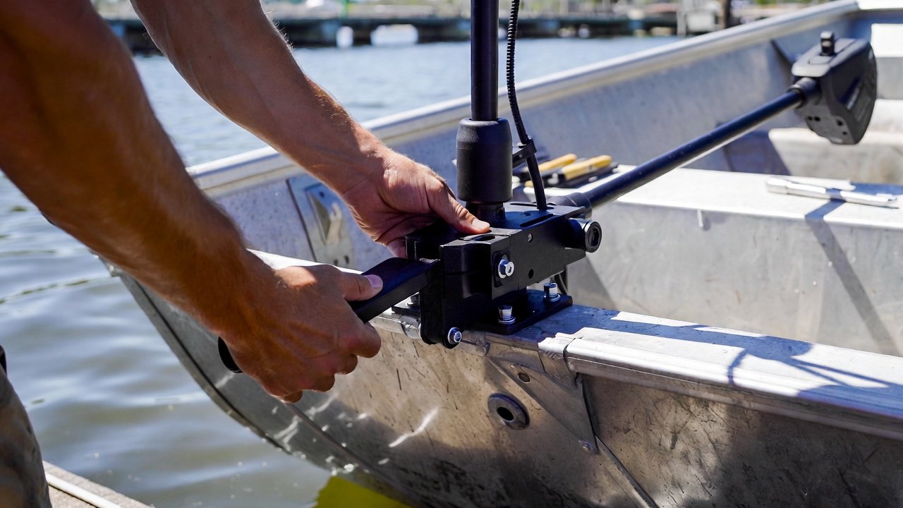 Close-up of hands mounting an electric trolling motor bracket onto the transom of a small aluminum jon boat at a dock